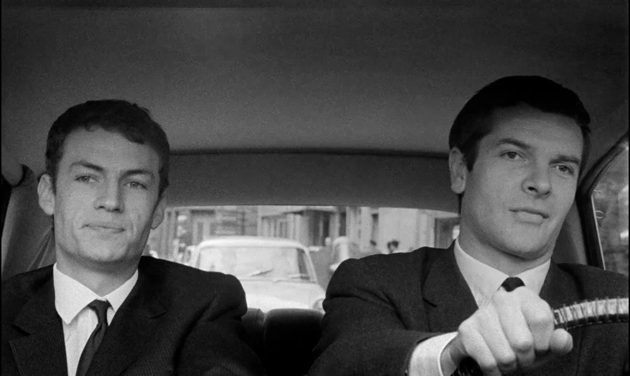 Bay of Angels (1963) movie still: medium — Two men in suits sit in the front seats of a moving vehicle. The car interior is framed against an…