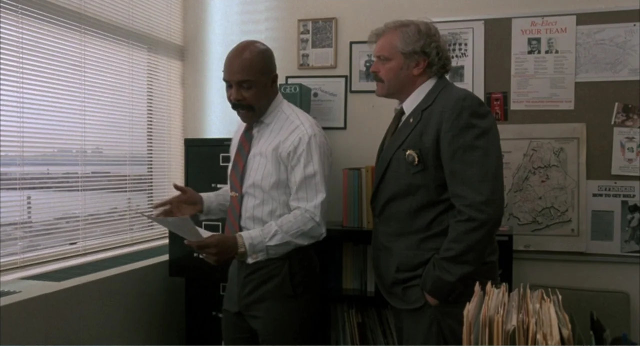 F/X (1986) movie still: medium — A cramped detective office features a bulletin board with posters and window blinds. Two men in dress shirts and…