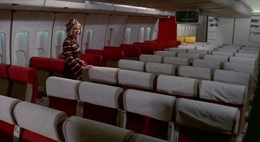 Lisa and The Devil (1973) movie still: wide — The interior of a passenger jet with rows of red and white seating. A woman stands in the aisle of the…