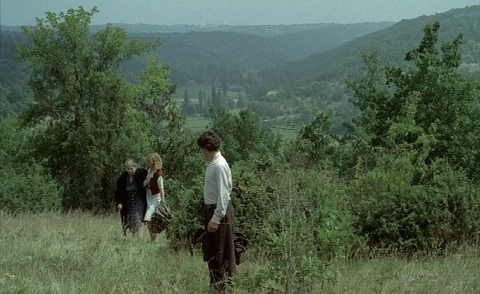 Lacombe, Lucien (1974) movie still: wide — A grassy hill overlooks a rolling valley covered in lush trees. Three people walk down the slope through…