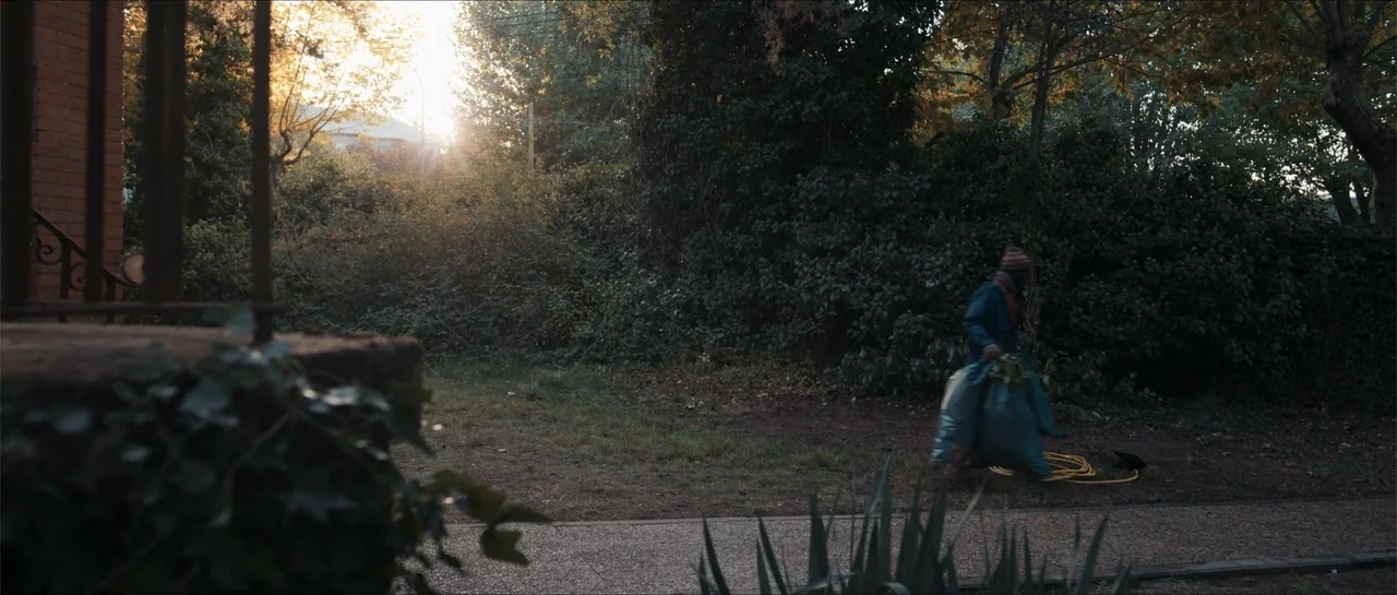 Historias lamentables (2021) movie still: wide — A residential front yard features dense foliage and a brick house porch side frame. A person in a…
