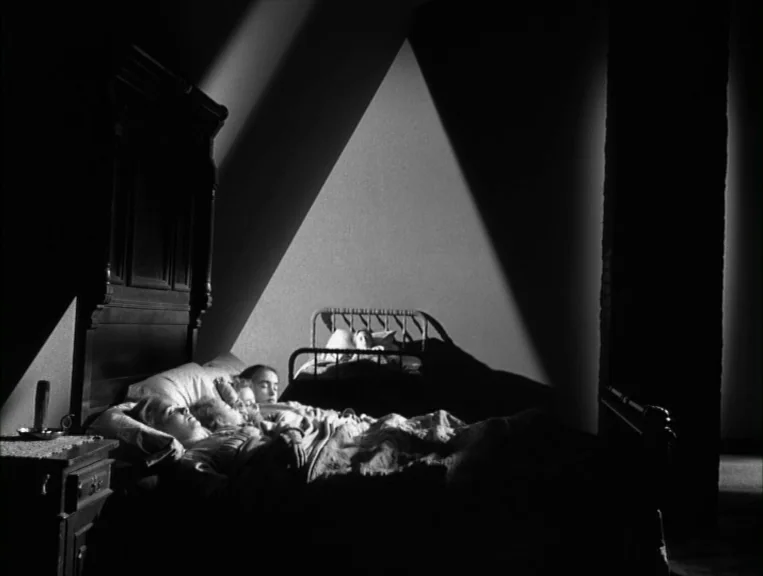 The Night Of The Hunter (1955) movie still: medium — A dark, sparsely furnished room contains two separate iron-framed beds arranged side-by-side.…