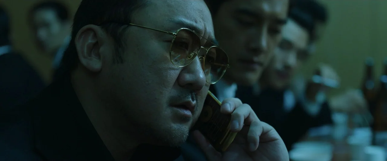 The Gangster, the Cop, the Devil (2019) movie still: closeup — An interior space with multiple men in dark suits seated at a table. A man in the…