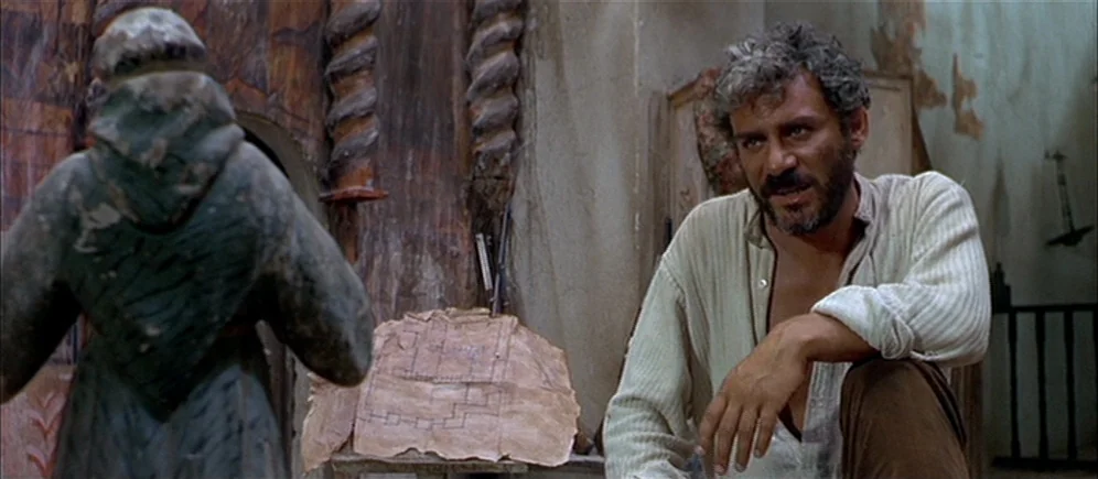 For A Few Dollars More (1965) movie still: medium — A dimly lit room with weathered stone walls and an old wooden carved statue. A middle-aged man…