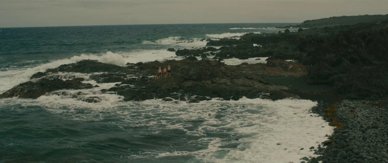 Evolution (2015) movie still: wide — A rugged volcanic shoreline features dark jagged rocks meeting the churning, deep green sea. Three small…