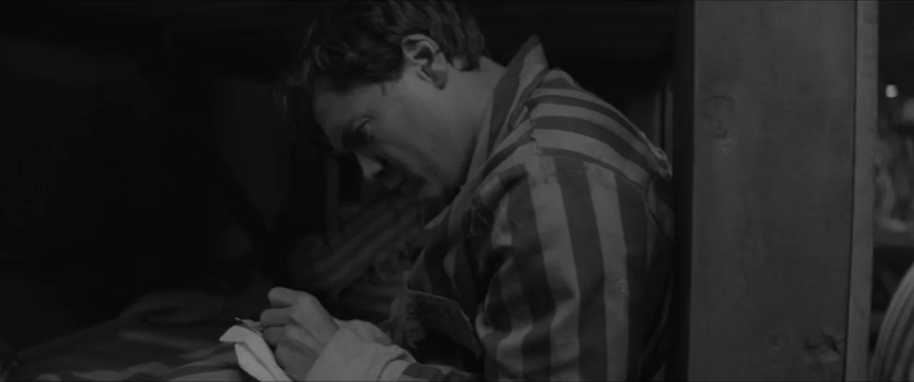 Bau, Artist at War (2025) movie still: medium — A confined, dark wooden bunk area contains a man in striped clothing. The man sits against a support…
