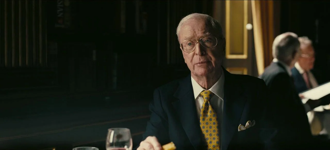 Tenet (2020) movie still: medium — An elderly man in a suit sits at a dark table in a dim restaurant. Other patrons appear blurry in the background…