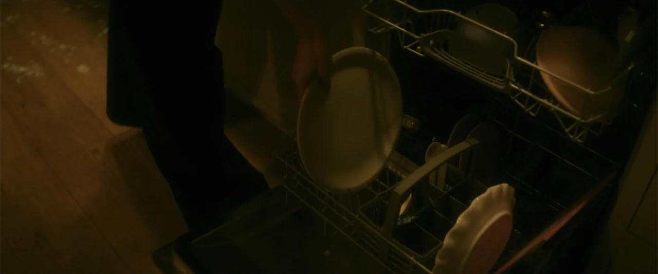 Black Bag (2025) movie still: closeup — A close view of an open dishwasher interior featuring metal racks and white ceramic plates. A person's hand…