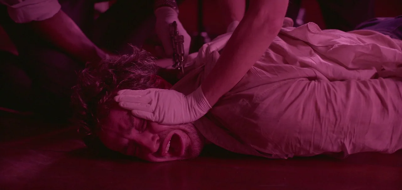 Foe (2023) movie still: medium — A man lies on a floor while being held down by another person. The captor presses a handheld syringe into the man's…