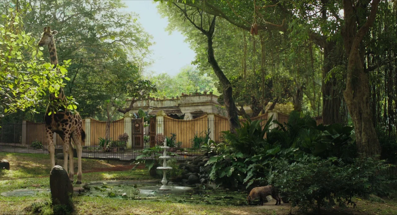 Life of Pi (2012) movie still: wide — A zoo enclosure features a small pond with a central fountain and a giraffe. A tan wooden wall separates the…