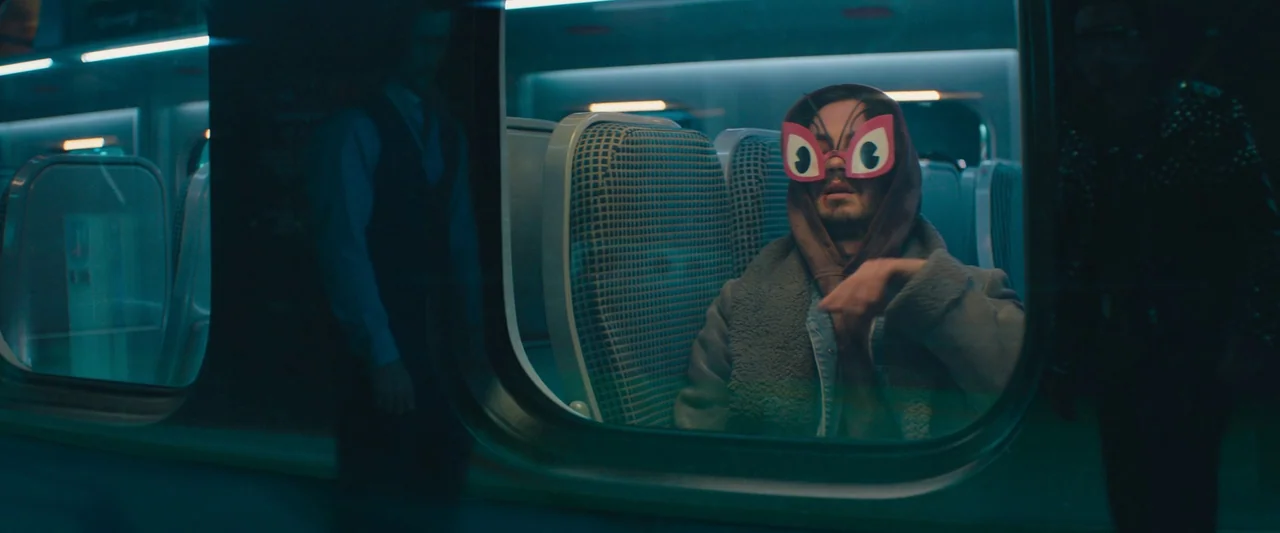Bullet Train (2022) movie still: medium — The interior of a commuter train at night with empty rows of seats. A man sits in a seat wearing a hooded…