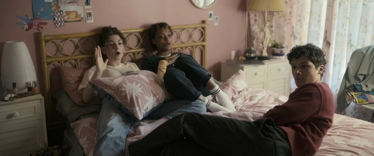 Talk to Me (2023) movie still: medium — A pink teenage bedroom features a bamboo headboard and floral patterned bedding. Three young people sit on…