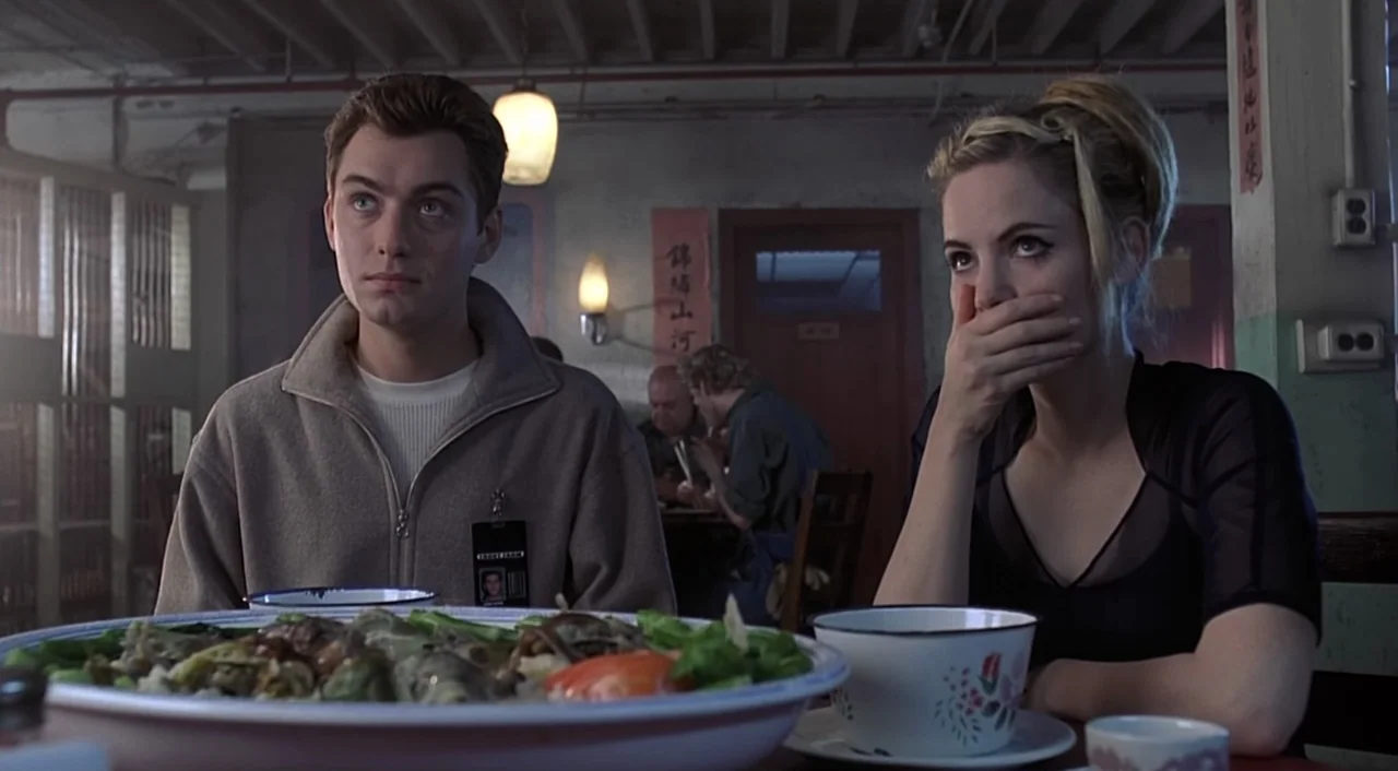 eXistenZ (1999) movie still: medium — A dimly lit restaurant with grey walls and Asian-style paper wall scrolls. A man and a woman sit at a table…