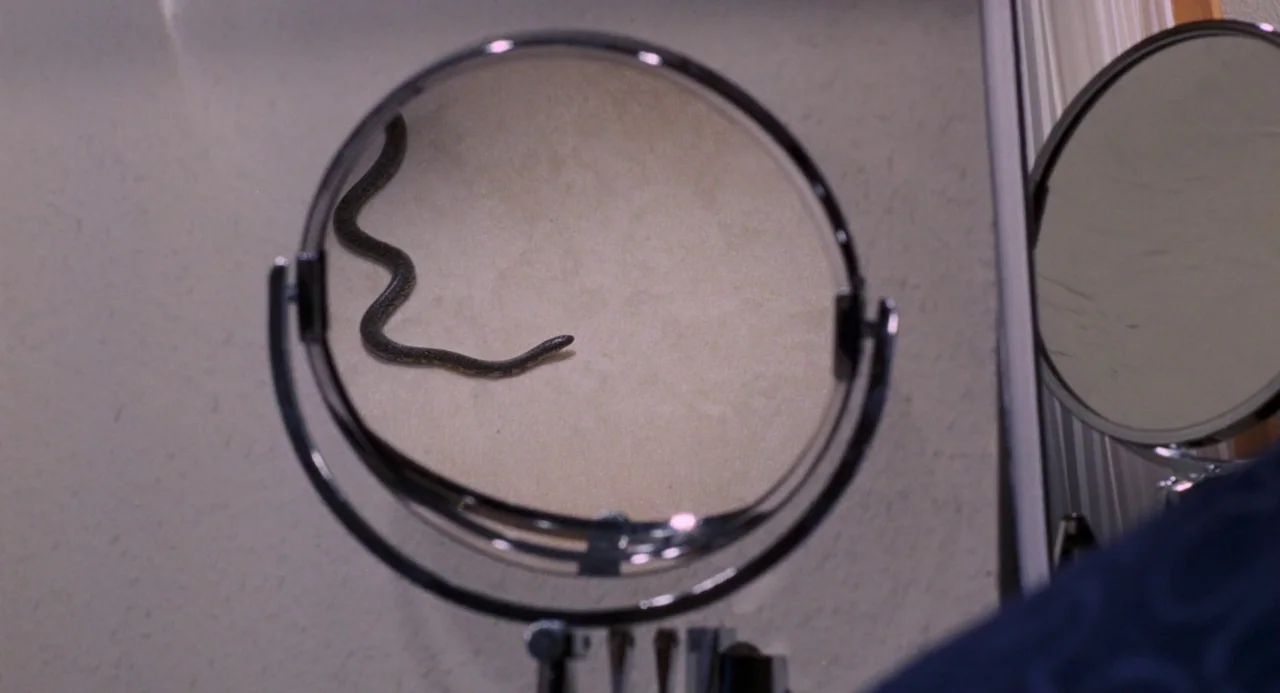 Live and Let Die (1973) movie still: closeup — A circular pedestal mirror sits on a surface, reflecting a wall. A thin snake travels across the face…