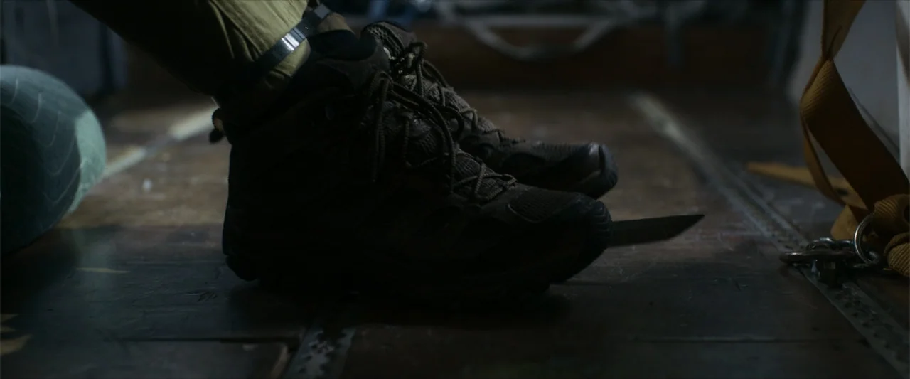 Flight Risk (2025) movie still: closeup — A dark, industrial cargo hold features a metallic floor with visible rivet lines. A pair of worn brown…