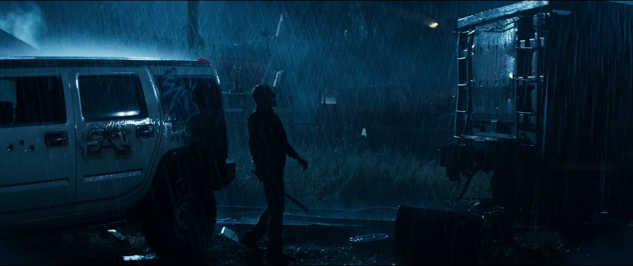 El Chicano (2018) movie still: wide — A dark, rain-drenched lot contains a white SUV and a large metallic trailer. A lone man walks between the…