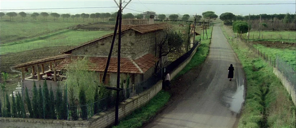 Deep Red (1975) movie still: wide — A rural Italian road stretches through green fields past a stone farmhouse with tiles. A person in a black coat…
