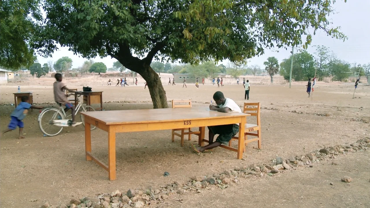 The Money Stone (2018) movie still: wide — An outdoor school yard features a large tree providing shade over wooden desks. A student sits at a desk…