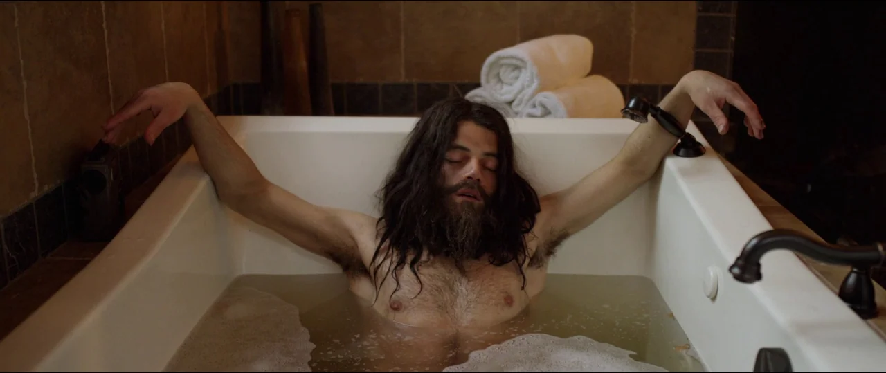 Buster’s Mal Heart (2016) movie still: medium — A man with long hair sits in a bathtub filled with murky water. He reclines with his arms draped…