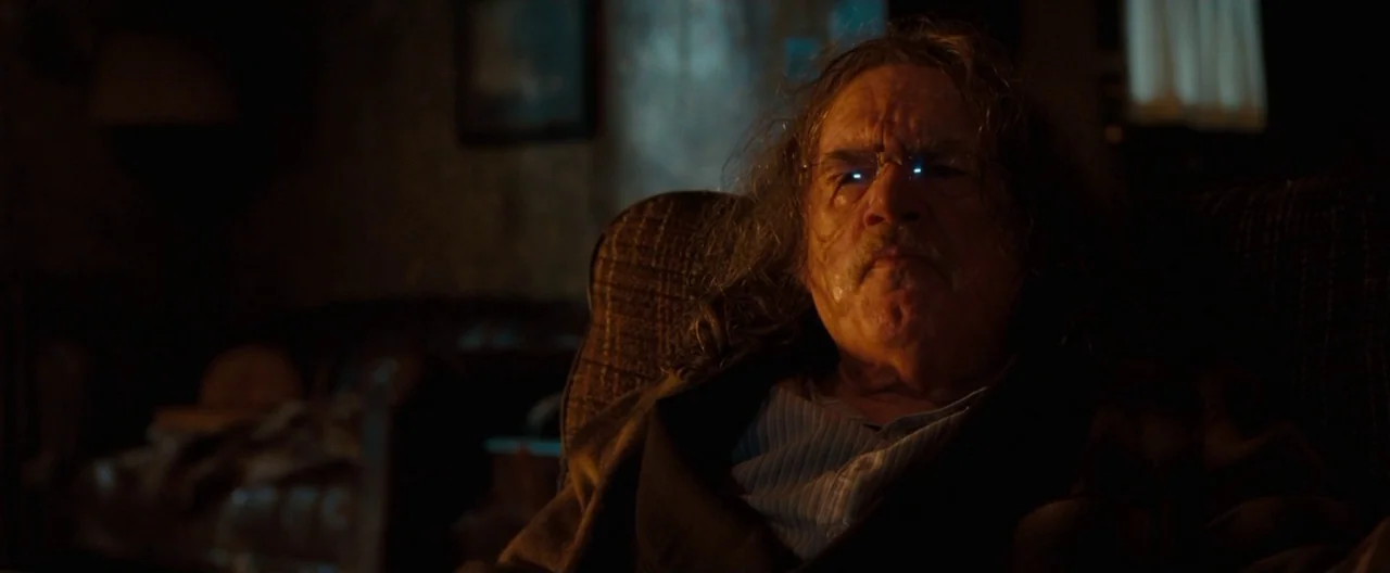 Trick ‘R Treat (2007) movie still: medium — A dimly lit room with muted wallpaper and indistinct furniture in the shadows. An elderly man sits in an…