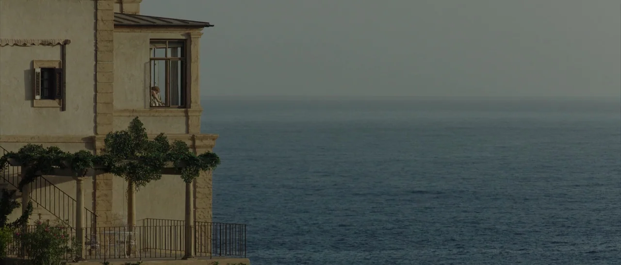 By The Sea (2015) movie still: wide — A stone villa perched on a cliffside overlooks a calm, expansive ocean during the day. A small figure is…