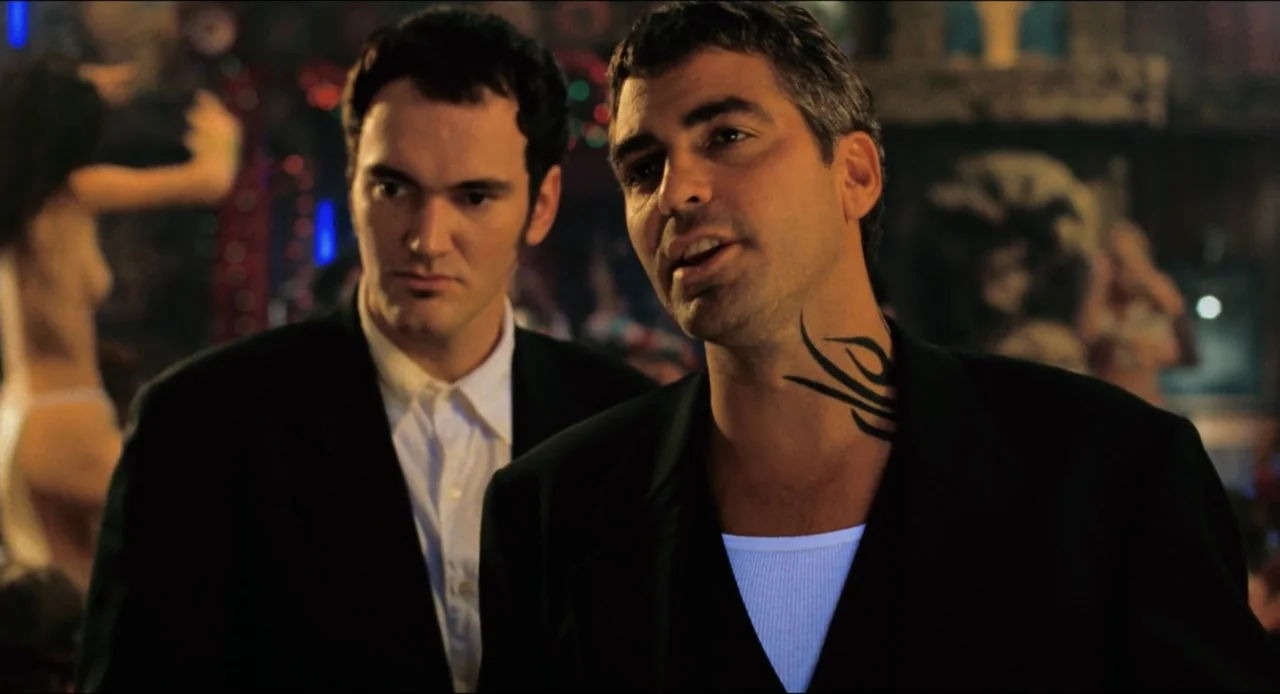 From Dusk Till Dawn (1996) movie still: medium — A dimly lit club interior features dark wood backgrounds with blurred figures. Two men stand in the…