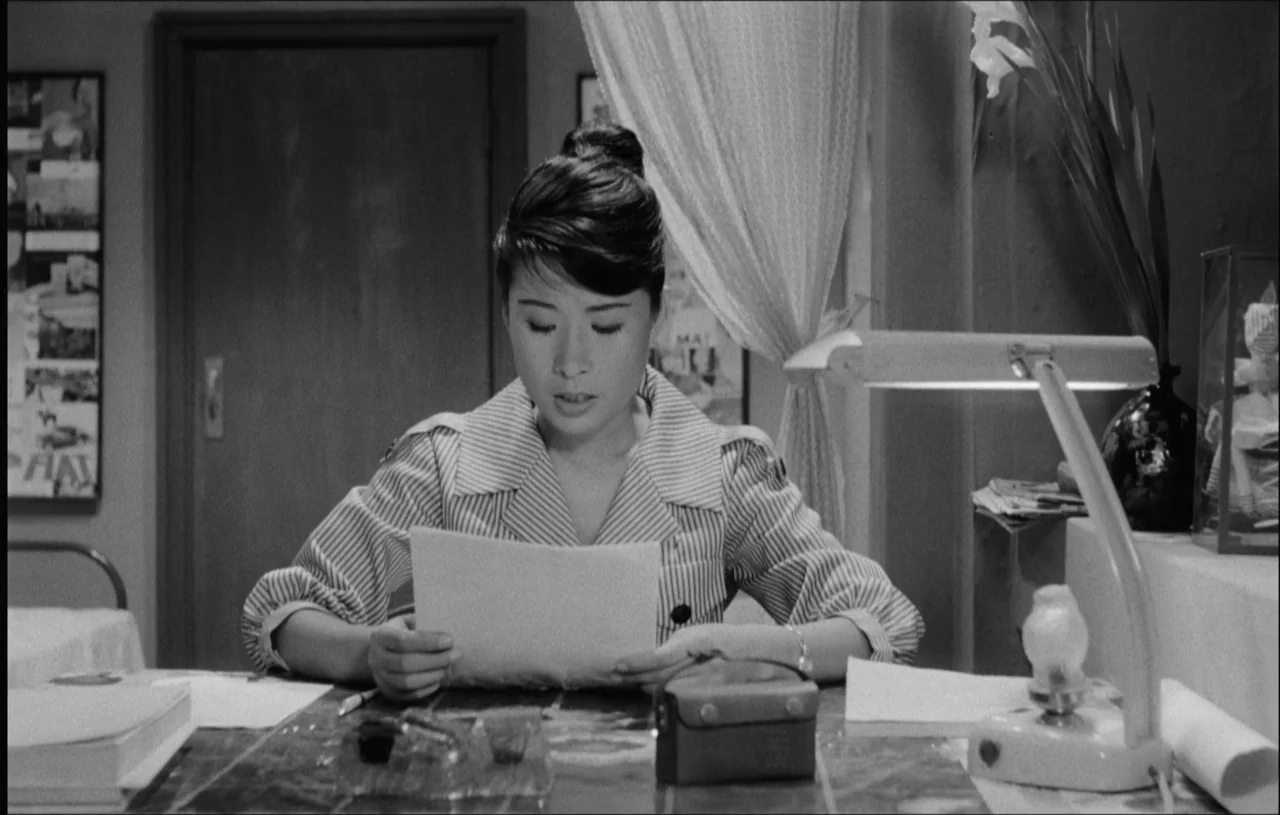 The Housemaid (1960) movie still: medium — An interior space with a desk lamp illuminating a woman reading a document. The background shows a door…