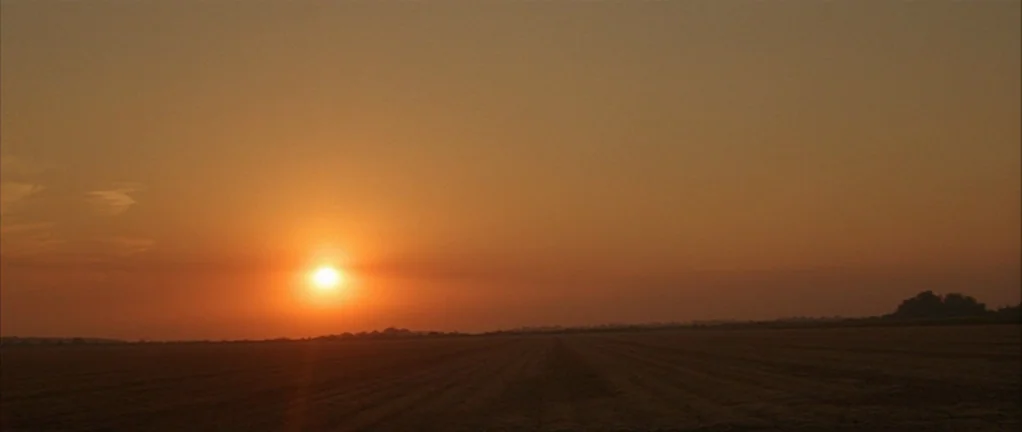 Shotgun Stories (2007) movie still: establishing — A flat, expansive agricultural field stretches to the horizon under a clear orange sky. The sun…