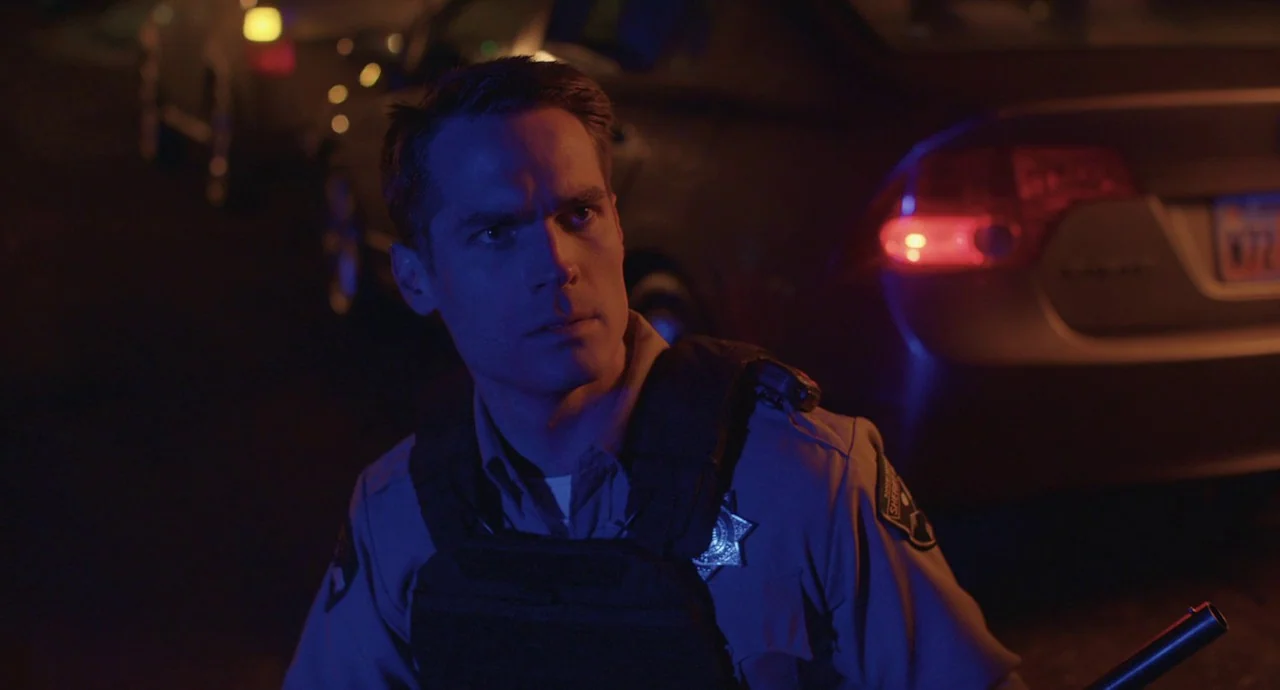The Wolf of Snow Hollow (2020) movie still: medium — A dark outdoor scene at night features a police officer in tactical gear. The officer watches…
