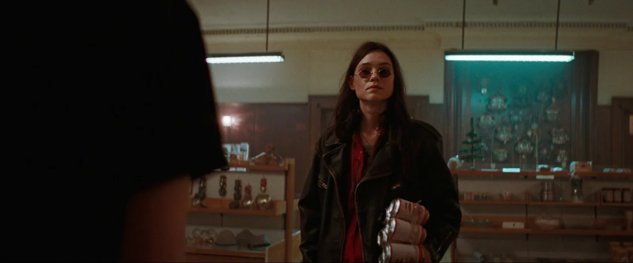 Cuckoo (2024) movie still: medium — A dimly lit convenience store has wooden shelving units filled with various small products. A woman in a leather…