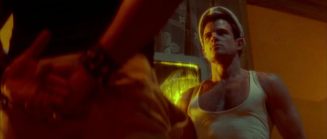 Querelle (1982) movie still: medium — A dimly lit room with lace curtains visible in the background against a window. A muscular man in a white tank…