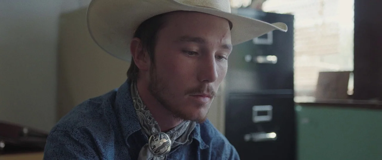 The Rider (2017) movie still: closeup — An interior space with a black metal filing cabinet visible in the blurred background. A young man wearing a…