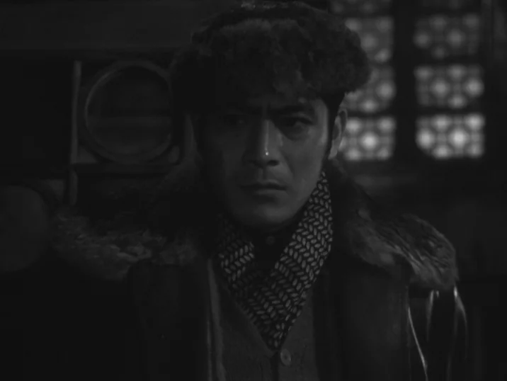 The Idiot (1951) movie still: closeup — A dark indoor space contains a man wearing winter clothing. He stares intently towards the side of the frame.