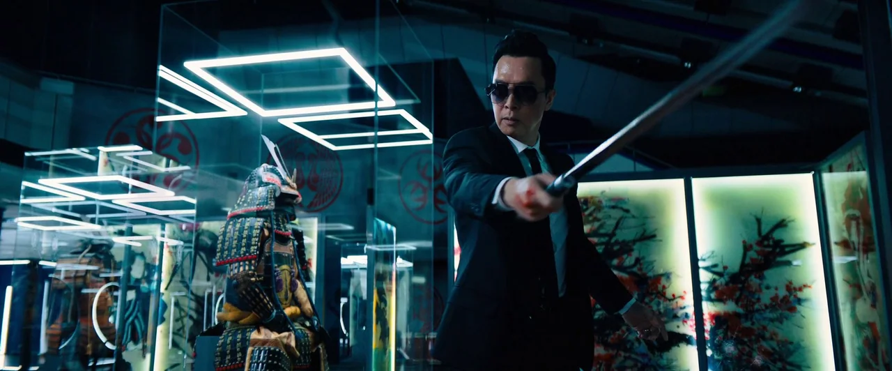 John Wick: Chapter 4 (2023) movie still: medium — A dark museum exhibit space features modern glass cases with lit square ceiling fixtures. A man in…