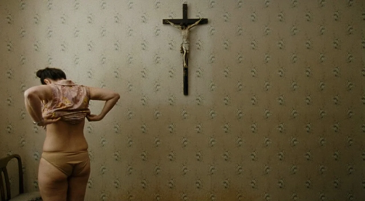 Paradise: Faith (2012) movie still: medium — An interior space features beige wallpaper with a repetitive floral pattern and a crucifix. A woman…