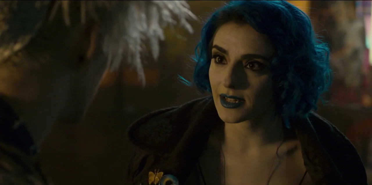 Mute (2018) movie still: closeup — A dimly lit space provides a dark backdrop for two people in conversation. A young woman with bright blue hair…