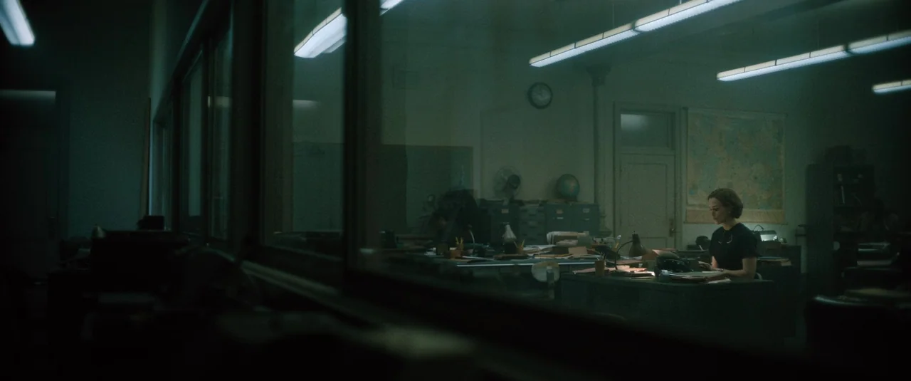 Boston Strangler (2023) movie still: wide — A dimly lit room functions as an office with a large glass window divider. A lone woman works at a desk…