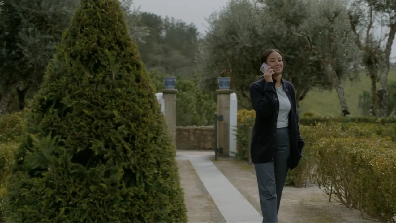 A Pinch of Portugal (2023) movie still: medium — A stone courtyard features manicured conical hedges and a central gravel pathway. A woman walks…