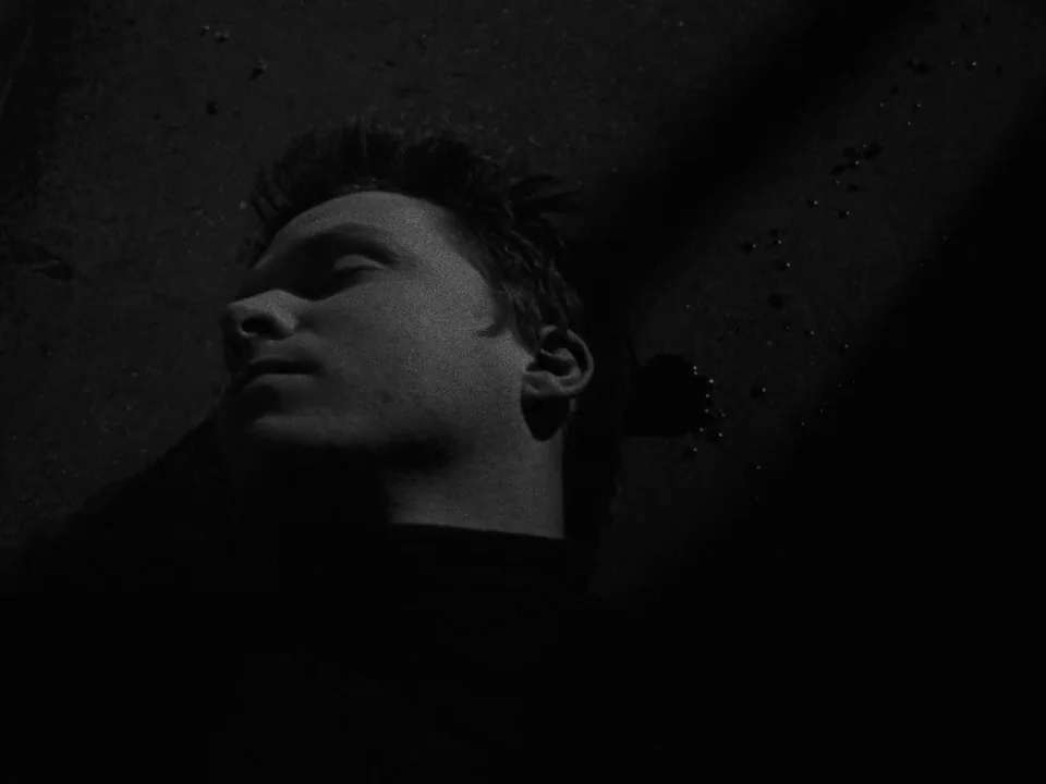 Following (1998) movie still: closeup — The head and shoulders of a man lie against a dark textured surface. Several small circular dark spots are…