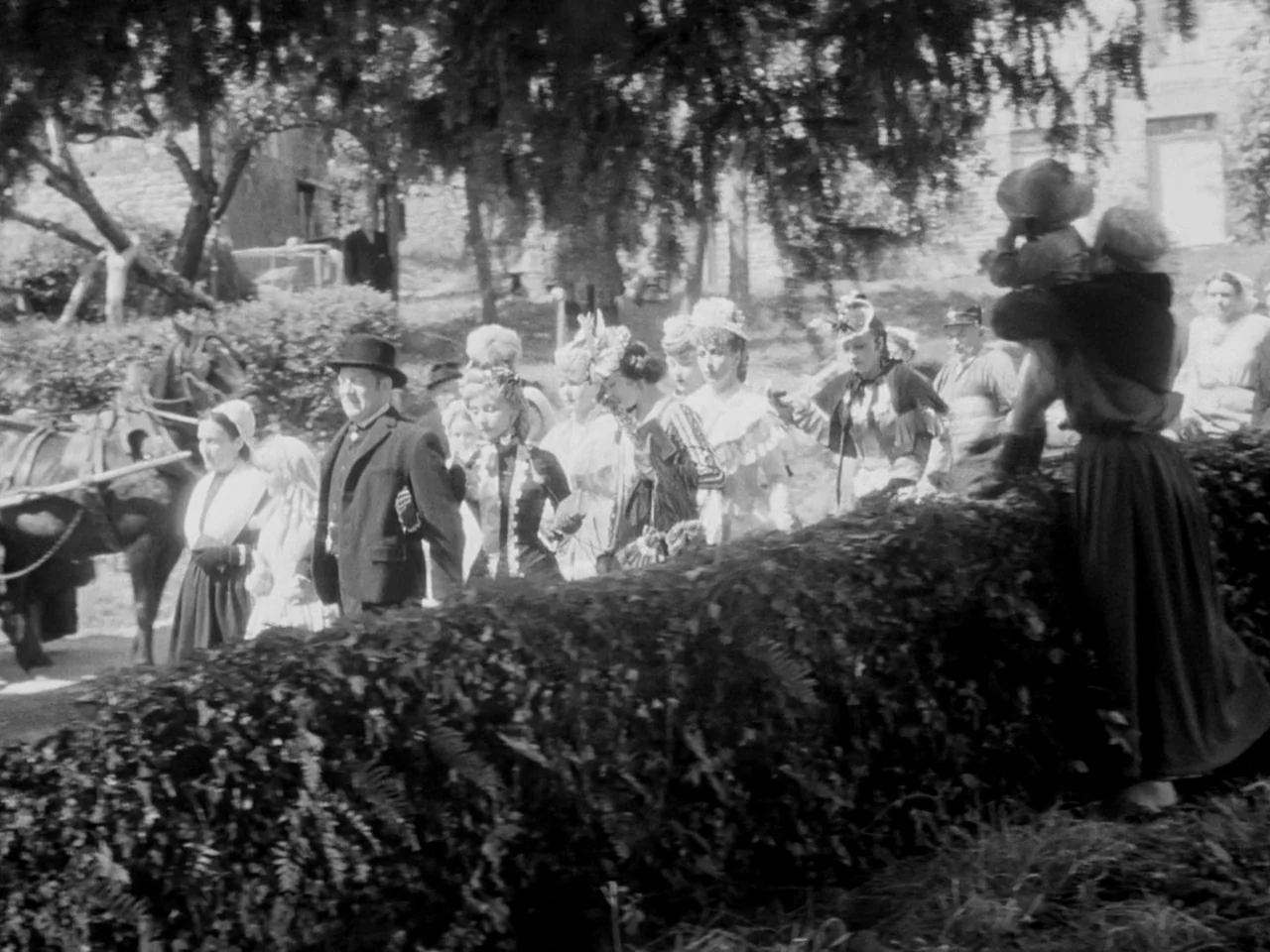 Le Plaisir (1952) movie still: wide — A group of people dressed in 19th-century formal clothing walks along a garden path. A person stands at the…