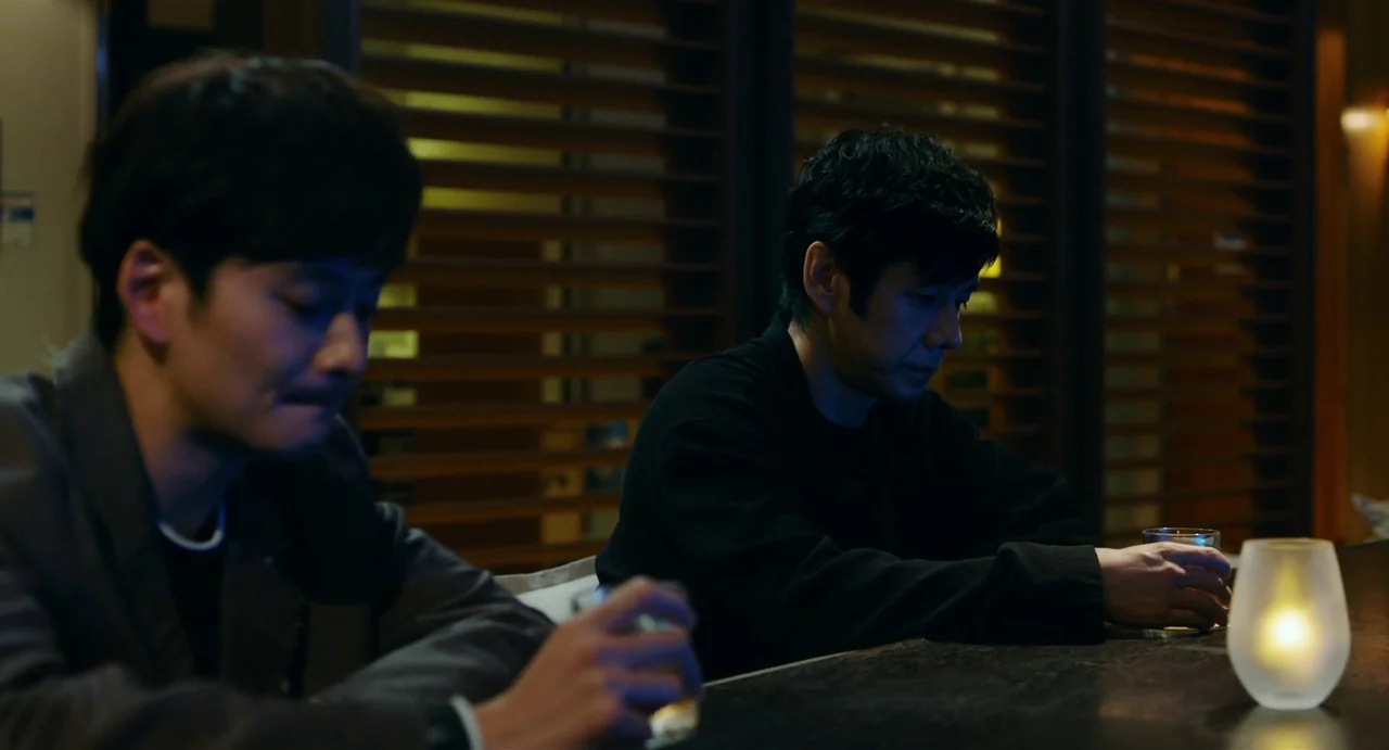 Drive My Car (2021) movie still: medium — Two men sit at a dark wooden bar counter with slatted wooden walls behind. A single lit glass candle…