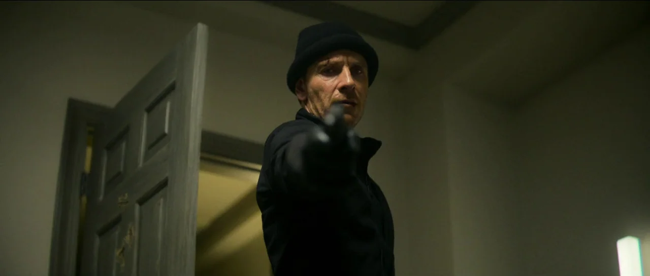 The Killer (2023) movie still: medium — A dark, dimly lit corridor features a partially open gray door. A man wearing a black beanie and jacket aims…