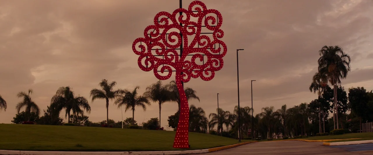 Stars At Noon (2022) movie still: establishing — An outdoor park area features a large, stylized tree sculpture covered in red LED lights. The scene…
