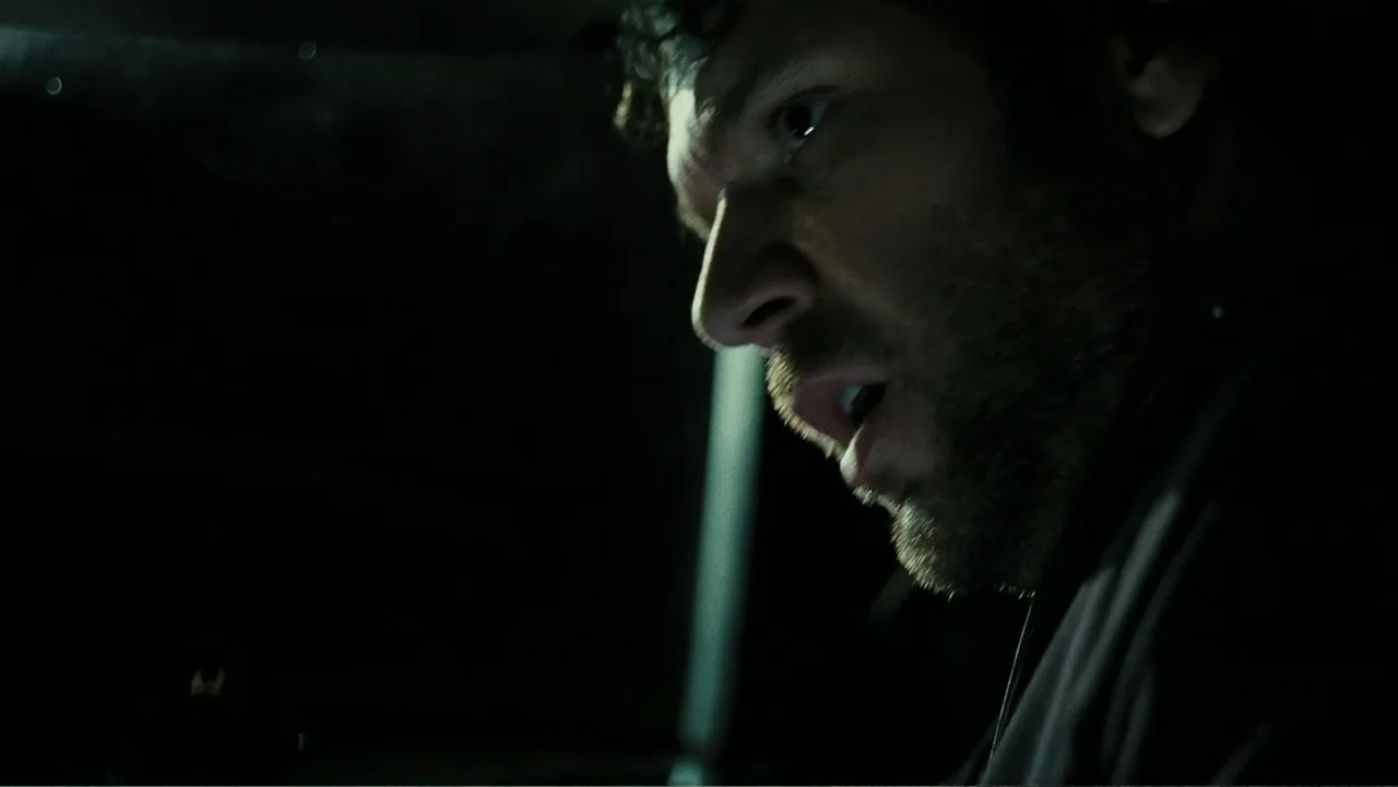 Mr. Brooks (2007) movie still: closeup — A dark, cramped interior space of a vehicle during the night. A man with dark stubble looks out the side…