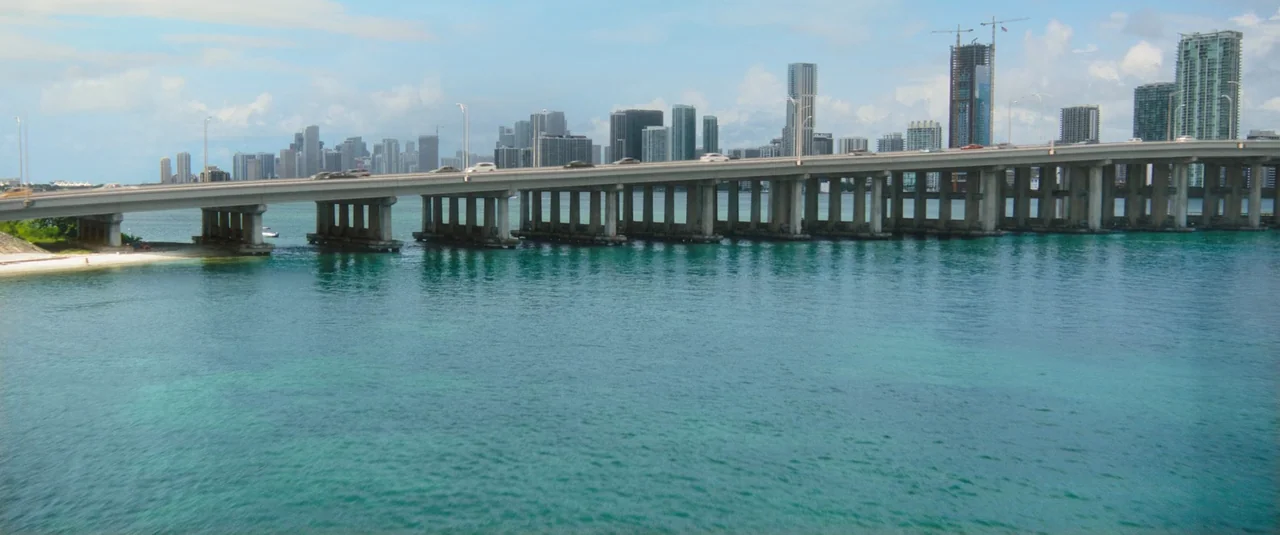 Do Revenge (2022) movie still: establishing — A long concrete bridge spans over a wide expanse of shallow turquoise water. The cityscape with…