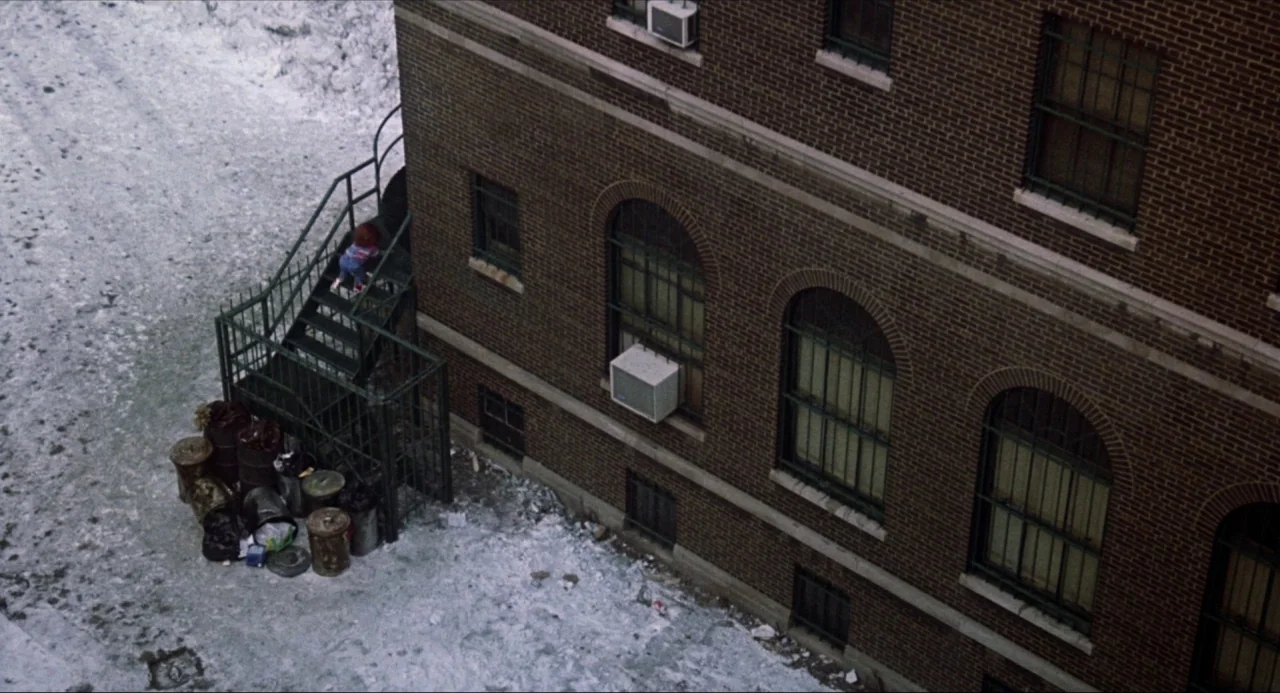 Child’s Play (1988) movie still: wide — A brick building exterior features an outdoor metal staircase surrounded by snow and trash. A small…