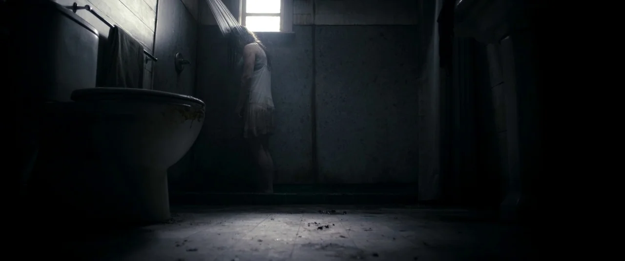 Evil Dead (2013) movie still: fullbody — A dilapidated bathroom with a dark, tiled floor and grime-streaked walls features a window. A woman stands…