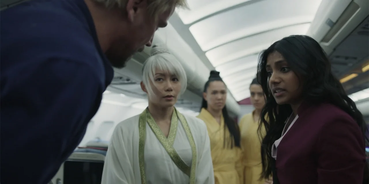 Fight or Flight (2025) movie still: medium — A group of people stand inside the narrow aisle of an airplane cabin. A woman in a maroon jacket faces…