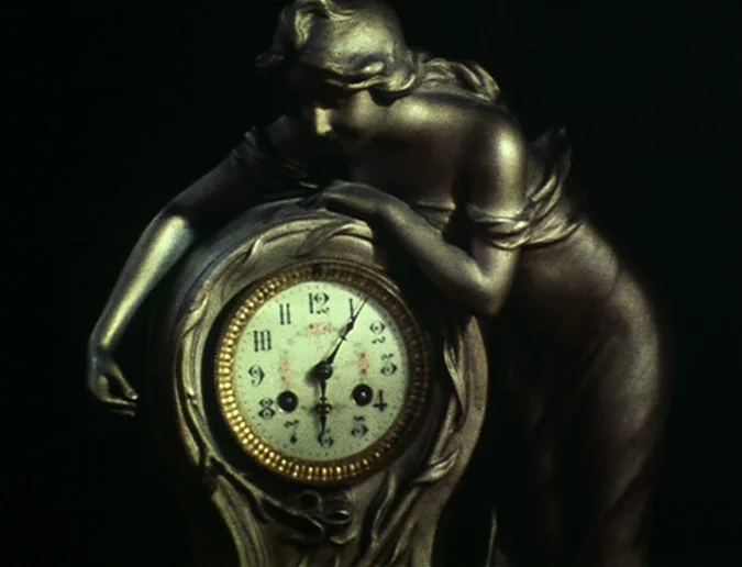 Daguerréotypes (1976) movie still: closeup — A bronze-colored figurine of a woman leans against an ornate analog mantle clock. The background is…