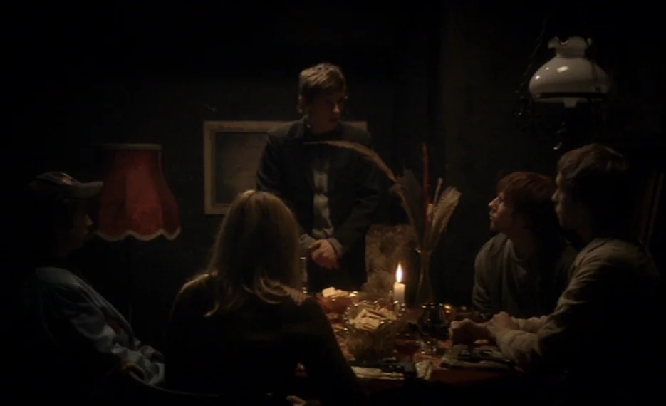 Dear Wendy (2005) movie still: medium — A dimly lit room features a small group of young adults seated at a table. A man stands behind the table…